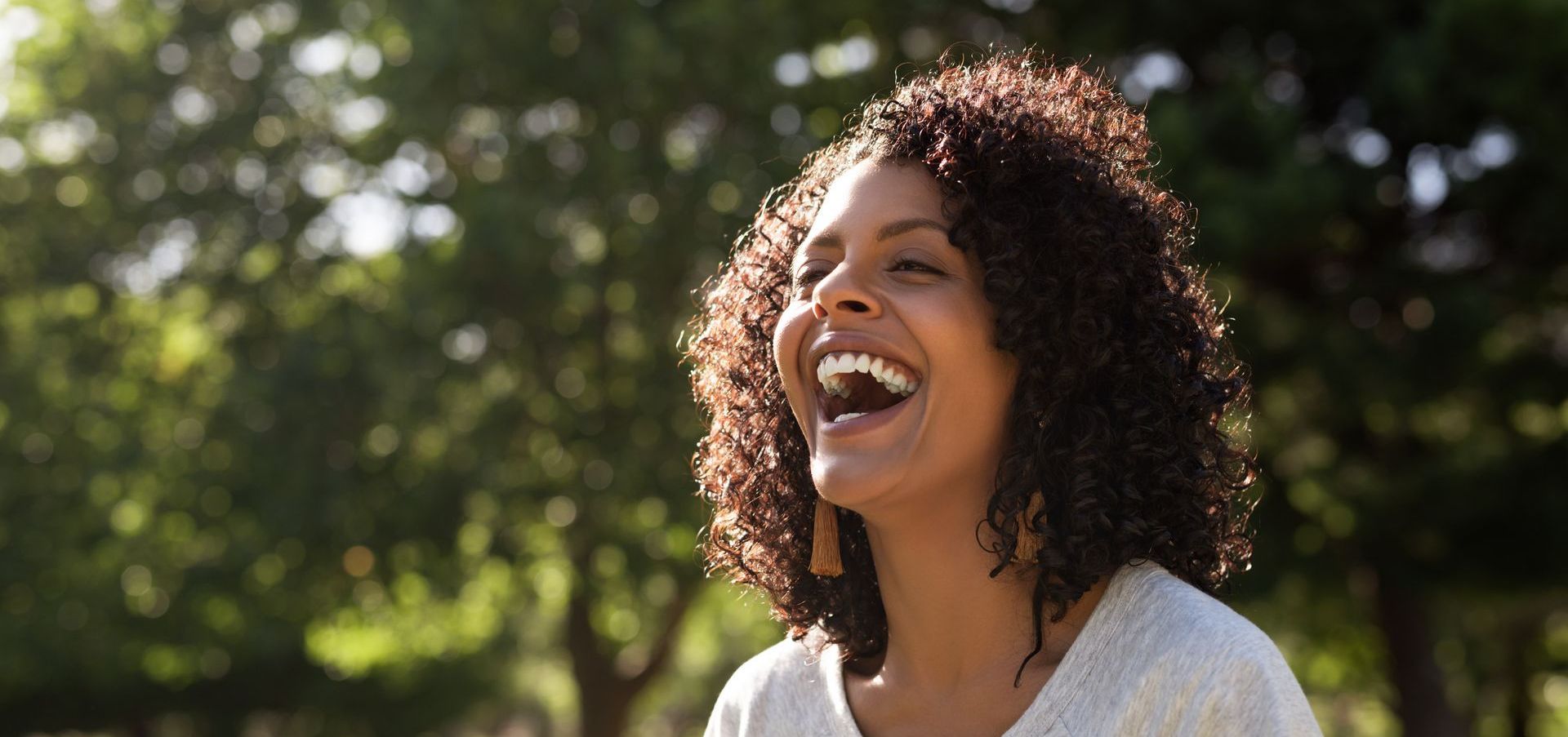 A woman with curly hair is laughing with her mouth open in a park.