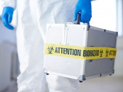 A person in a protective suit is holding a box with a yellow tape that says attention biohazard.