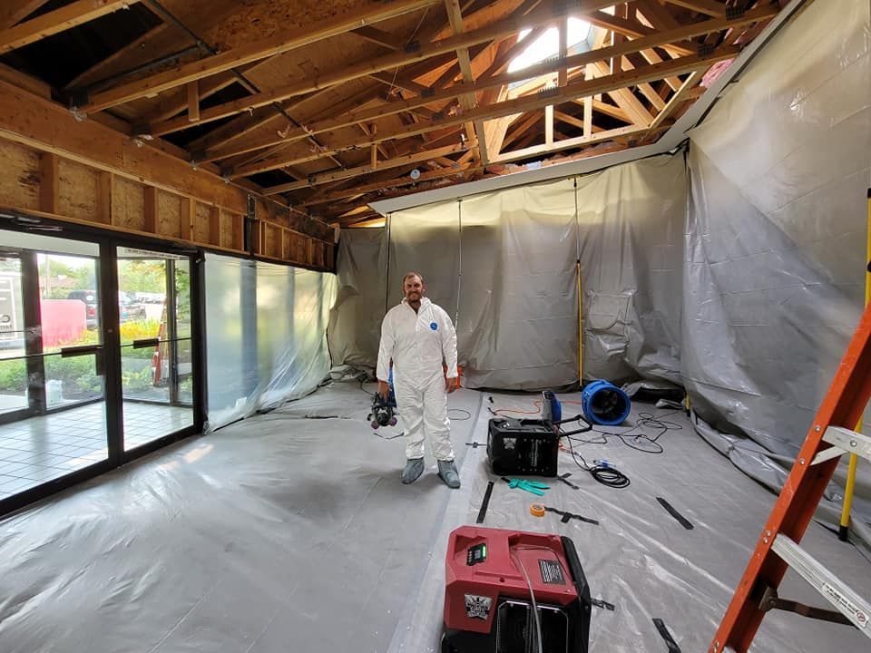 Man in protective suit stands in room under renovation; plastic sheeting on walls/floor, exposed beams, door on left.