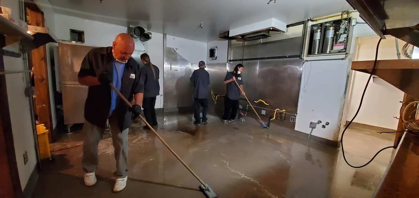 Four people sweeping water on a concrete floor in a commercial kitchen. Stainless steel surfaces and equipment are visible.