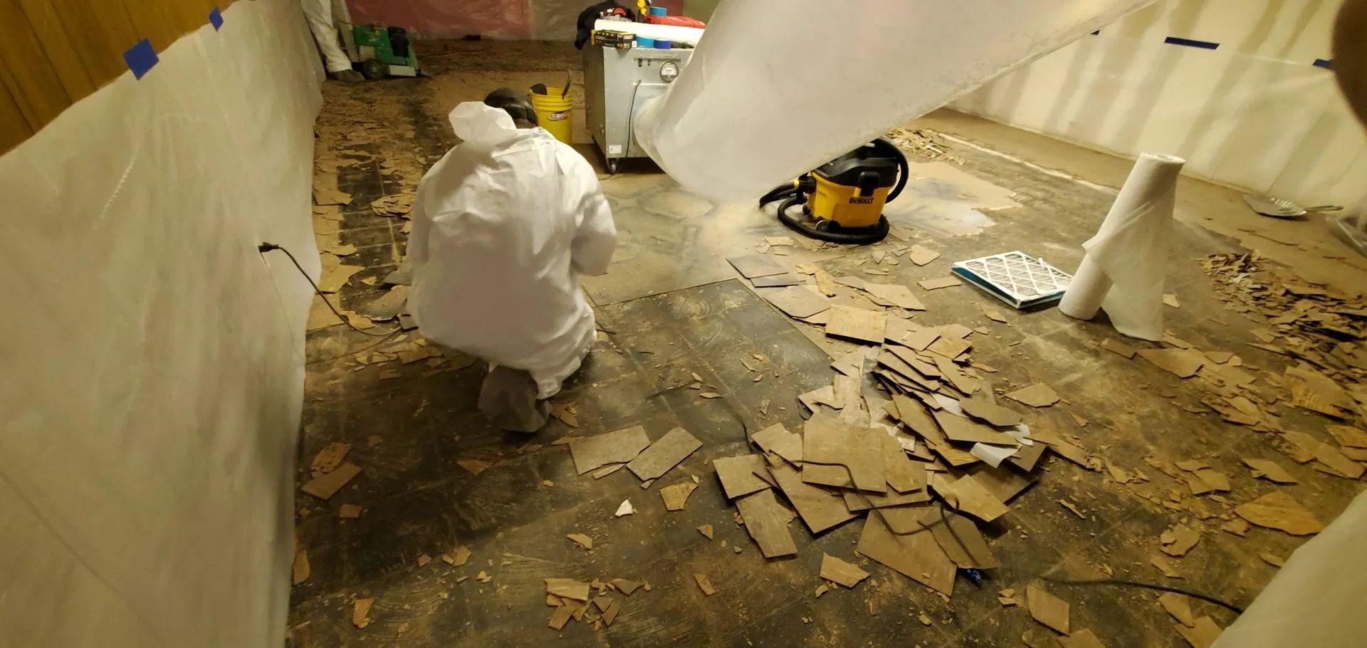 Person in protective suit working on a floor with debris, vacuum, plastic sheeting. Indoor setting.