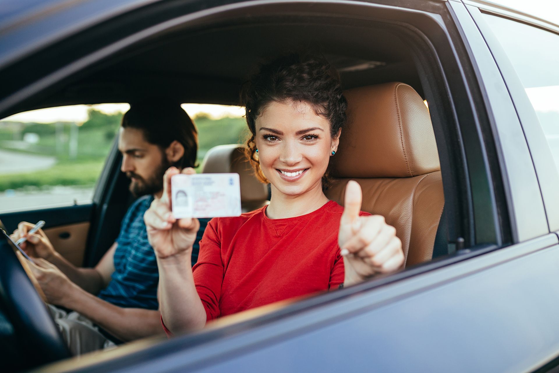 A woman is sitting in a car holding a driving license and giving a thumbs up.