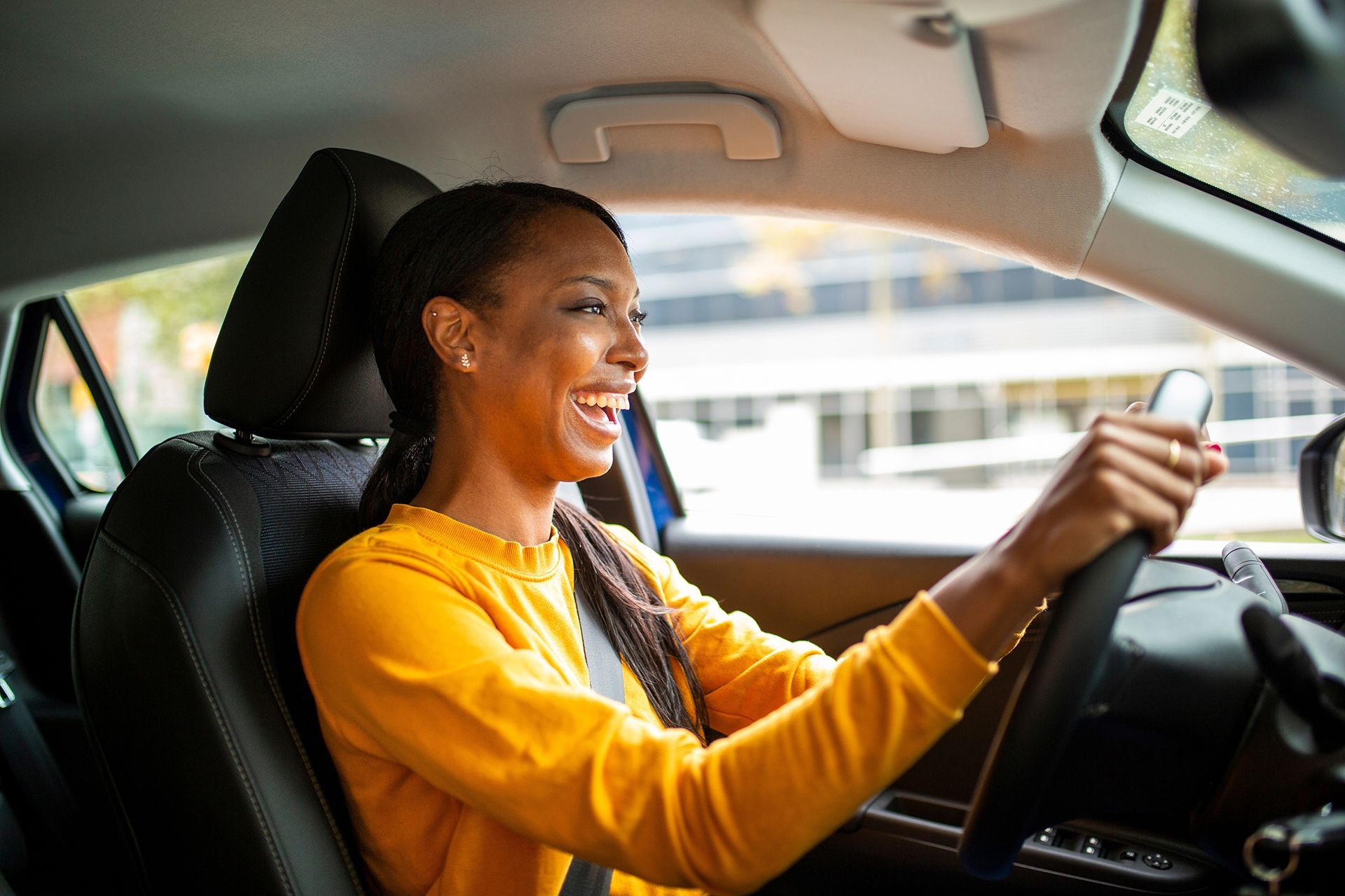 A woman in a yellow sweater is driving a car and smiling.