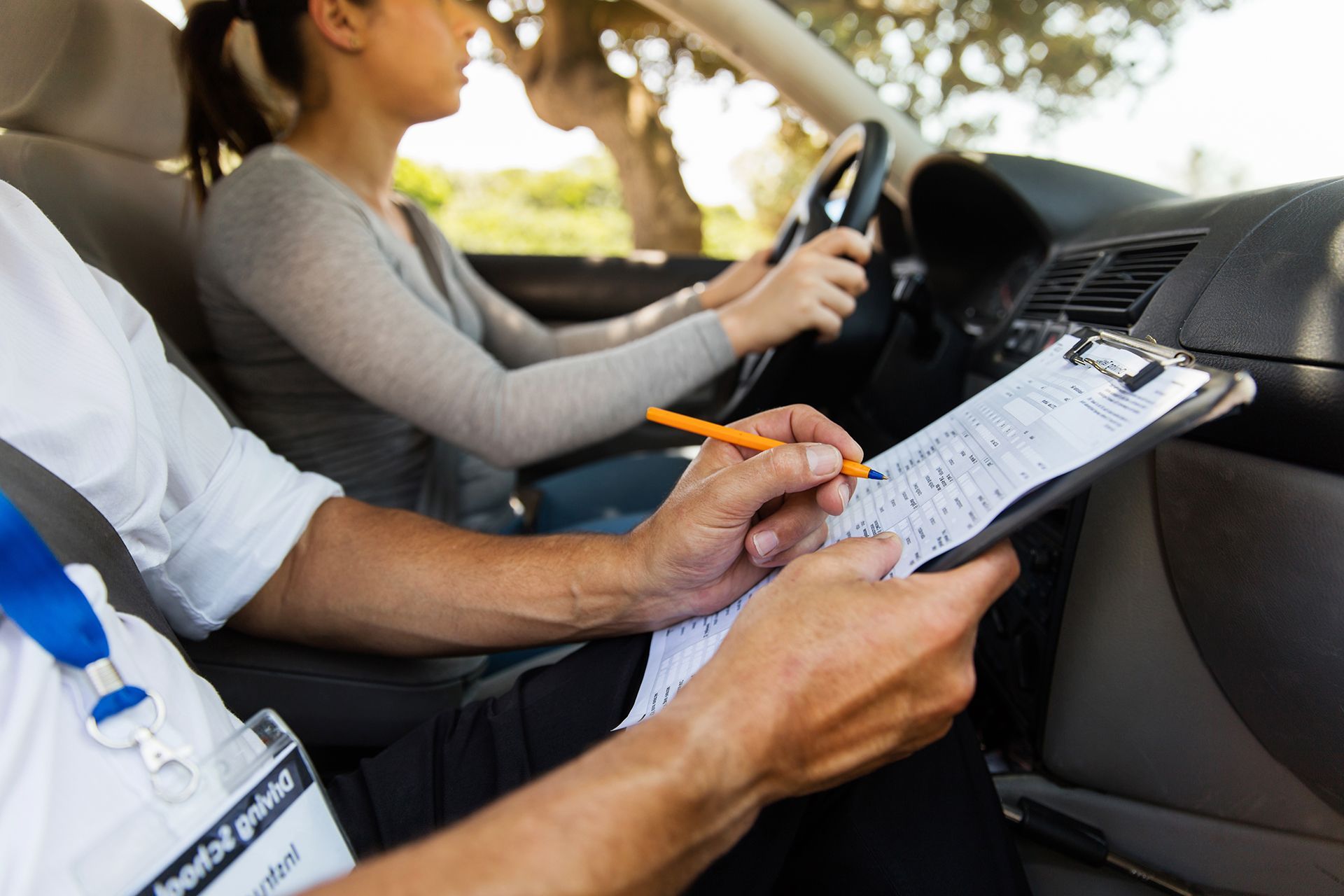 A woman is taking a driving test in a car while a man writes on a clipboard.