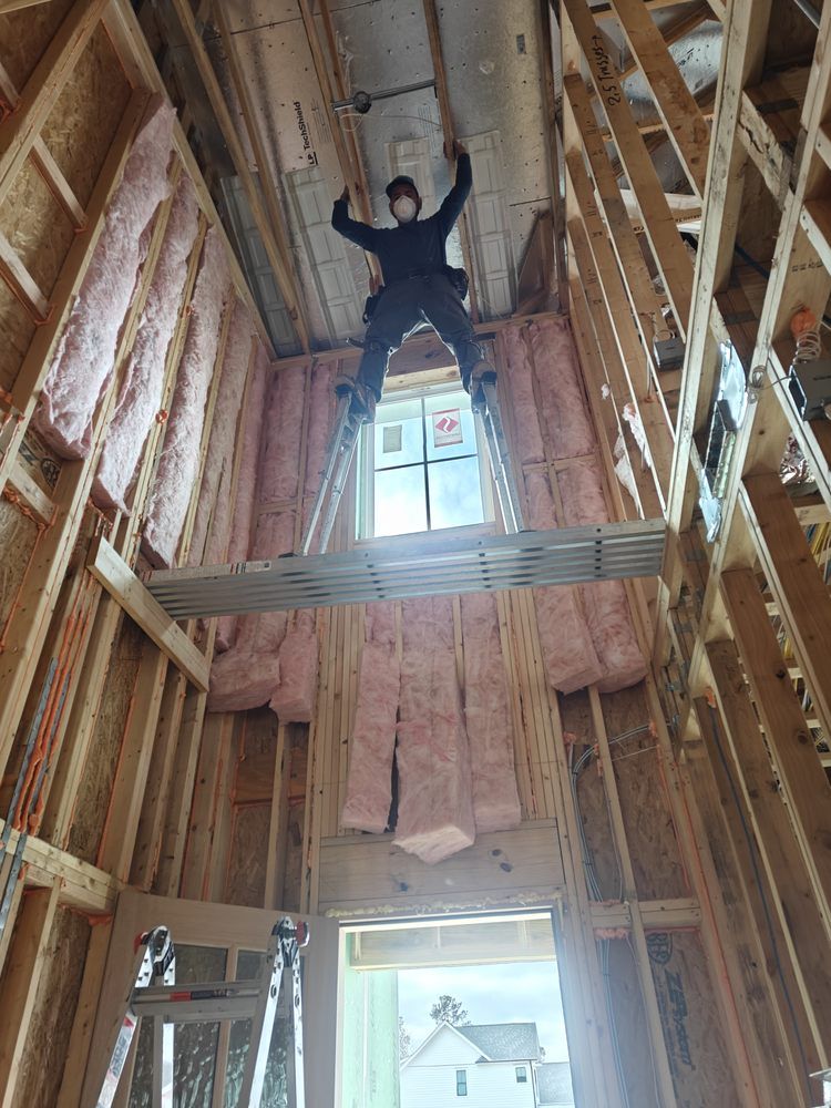 A construction worker on stilts works on the ceiling of a high-ceilinged, unfinished room framed with wood and insulation.