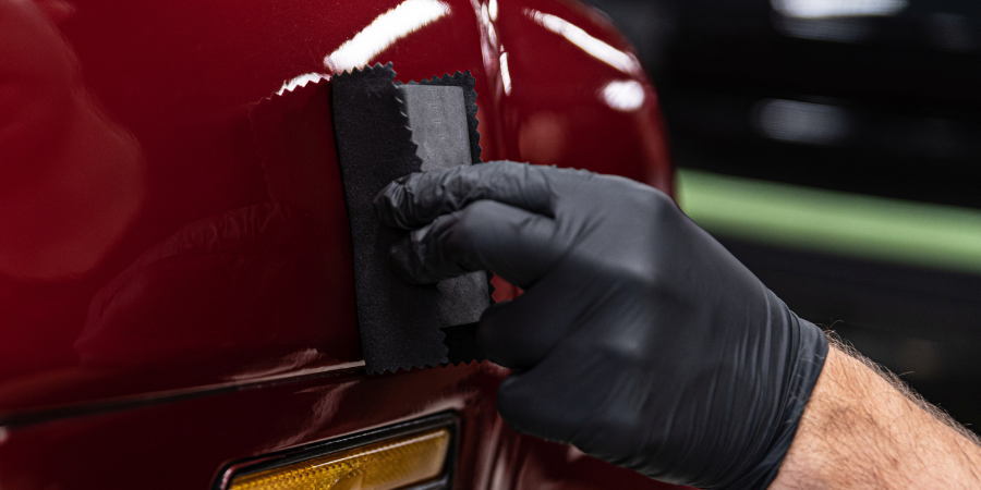 A person wearing black gloves is polishing a red car with a sponge.