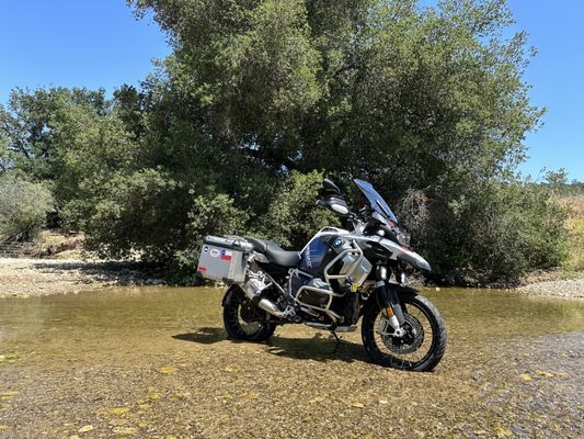 A motorcycle is parked on the side of a river next to a tree.