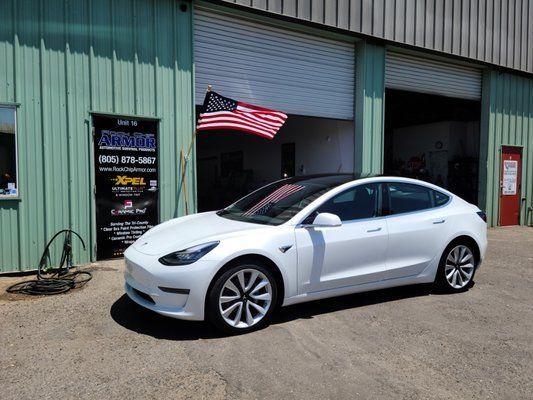 A white tesla model 3 is parked in front of a garage.