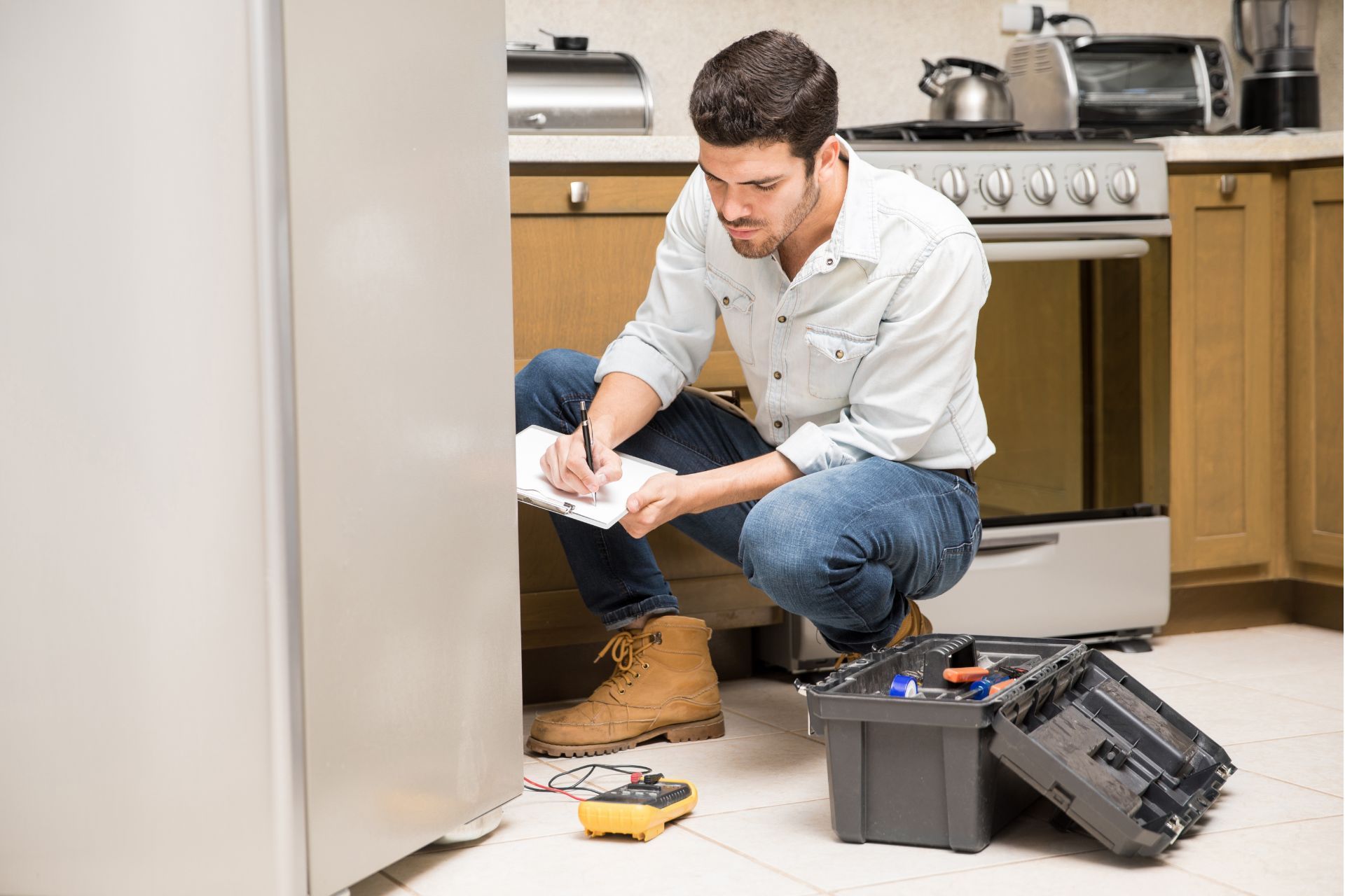 Man inspecting a refrigerator, squatting and writing on a notepad in a kitchen, tools nearby. Home Repair Services in Mt. Juliet TN
