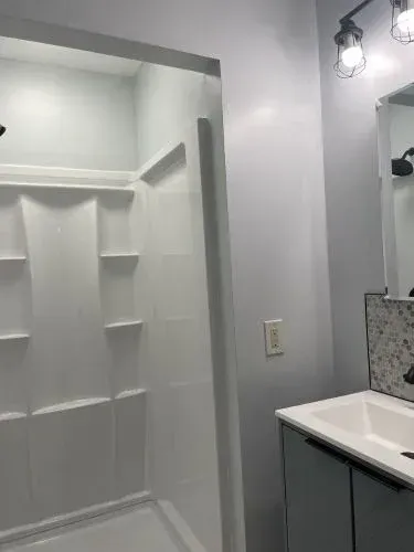 White bathroom with a built-in shower niche, vanity, and mirror under wall sconces.