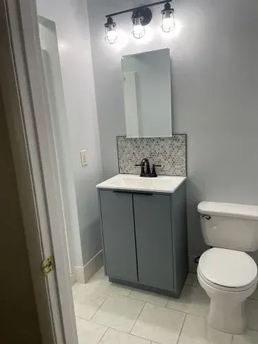 Small bathroom with gray vanity, patterned backsplash, toilet, and wall-mounted light fixture.