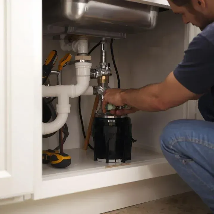 Man installing a garbage disposal under a kitchen sink, tools visible, indoors.
