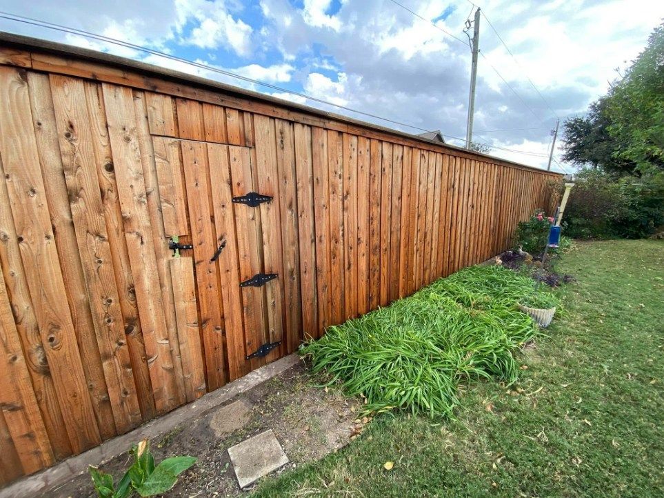 Wooden fence with a small door, surrounded by grass and plants under a cloudy sky.