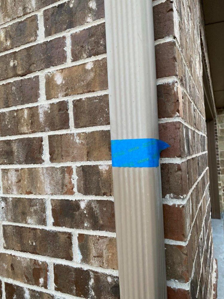 Beige rain gutter taped to a brick wall with blue painter's tape.