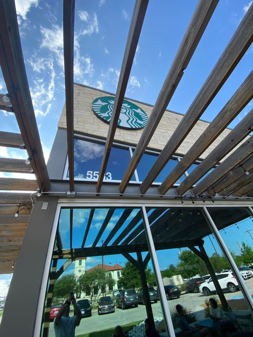 Starbucks building with large logo under a wooden pergola, reflecting blue sky and parking lot.