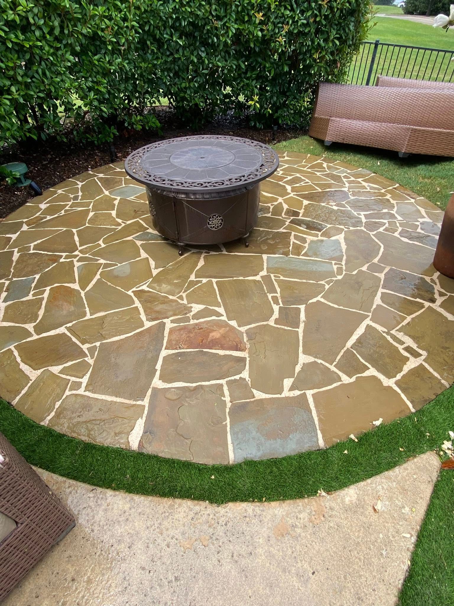 Round stone patio with fire pit, bordered by green grass and shrubs.