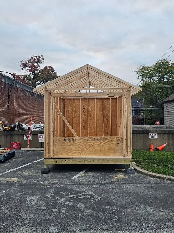 Wooden shed under construction in a parking lot, with a partially framed roof and walls. Cloudy sky.