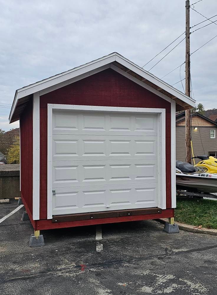 Red shed with white garage door, sitting on concrete blocks, parked on asphalt.