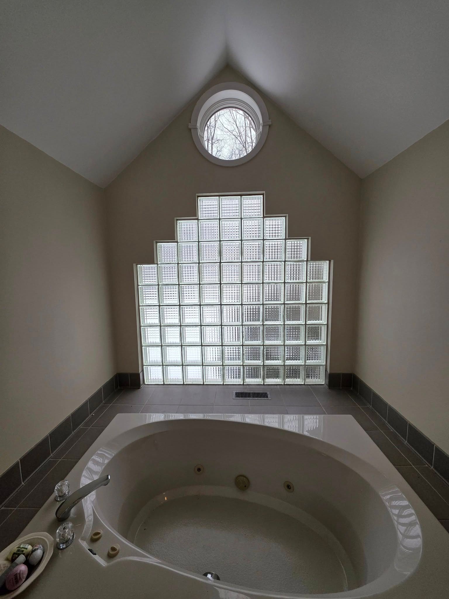 Bathroom with a white jacuzzi tub, glass block window, and circular window at the peak of the angled ceiling.