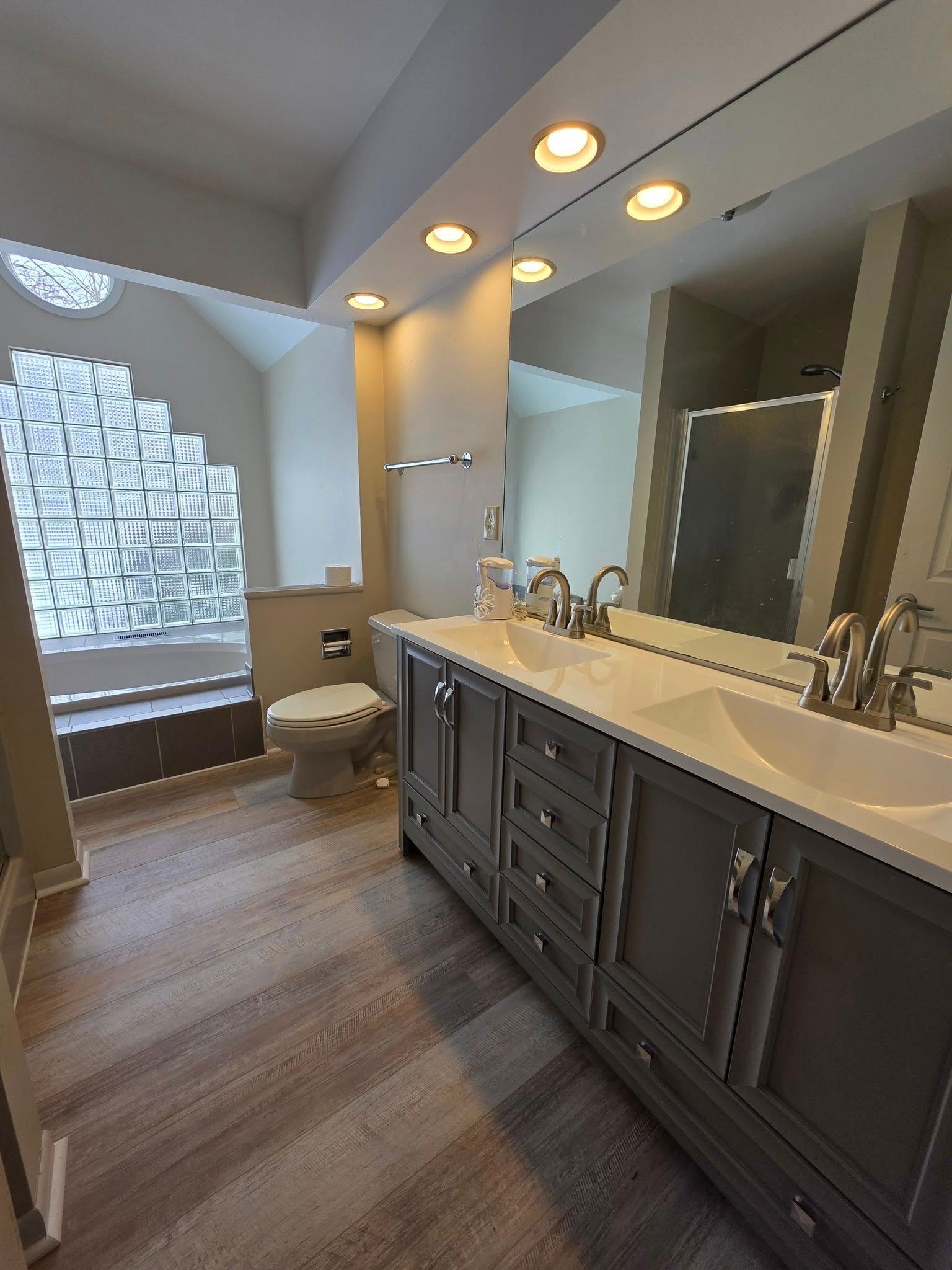 Bathroom with gray double vanity, large mirror, glass block window, and wood-look flooring.