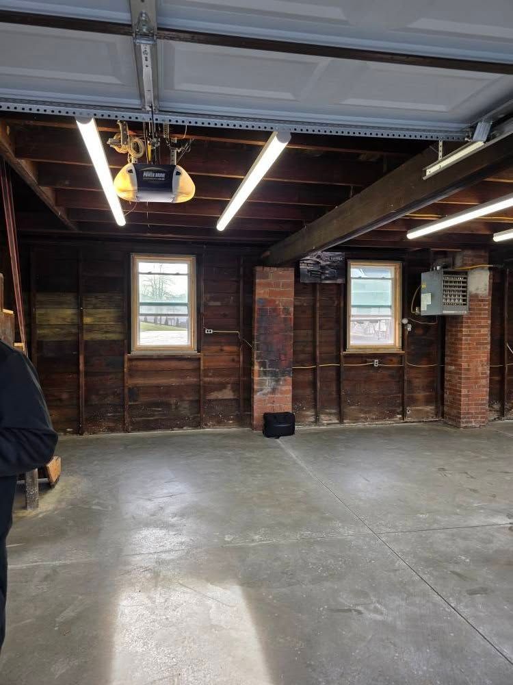 Empty garage interior with exposed wooden walls, concrete floor, two windows, and fluorescent lights.