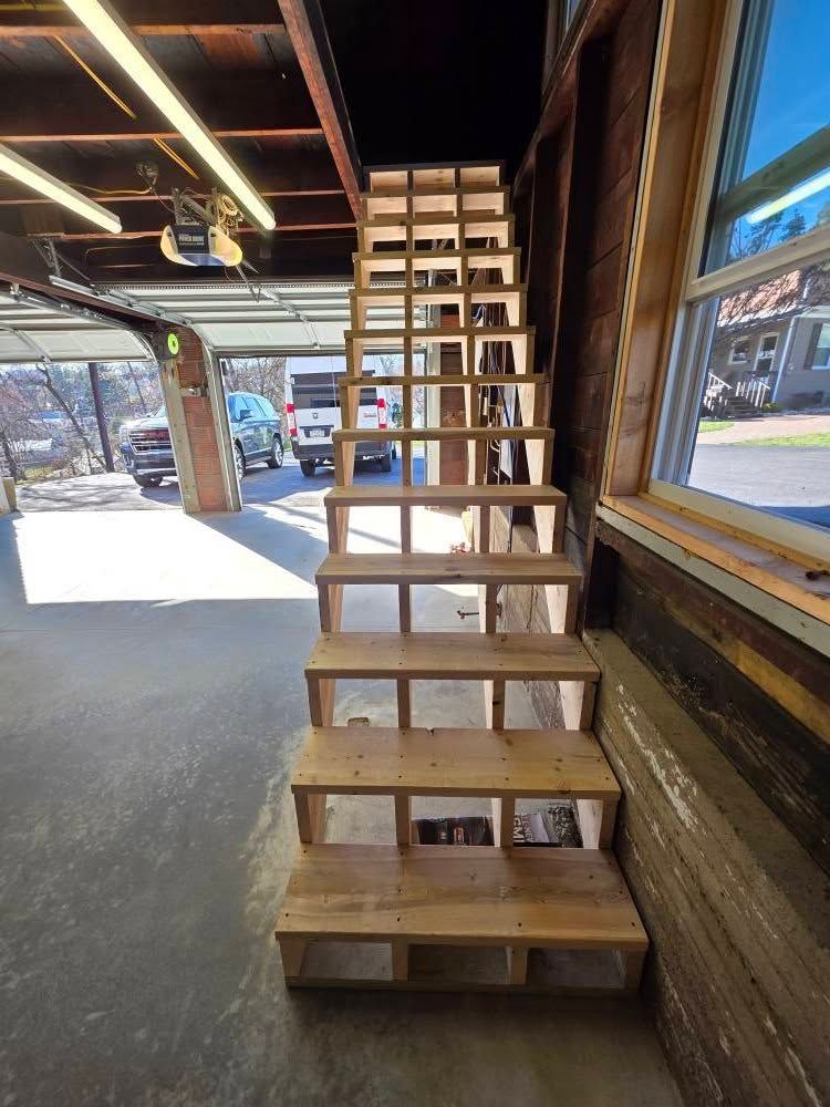 Wooden staircase inside a garage. Stairway leads upwards, with an open-riser design. Sunlight floods the interior.