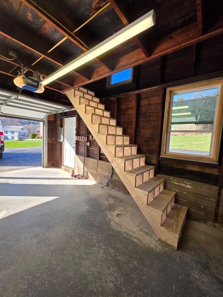 Wooden staircase built in a garage, leading upwards. Sunlight streams through an open door and window.