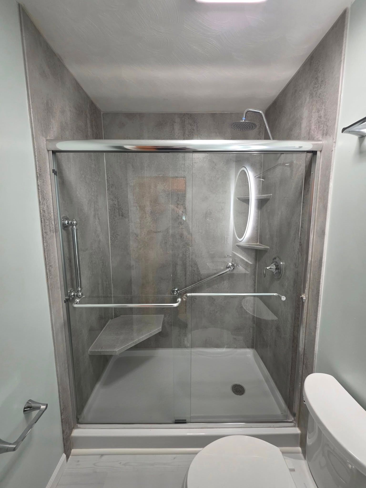 Accessible shower with bench, grab bars, and glass doors; gray tile walls, chrome fixtures.