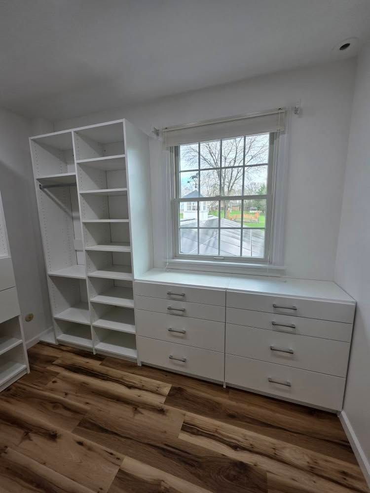 Built-in white closet with drawers and shelving beside a window in a room with wood flooring.