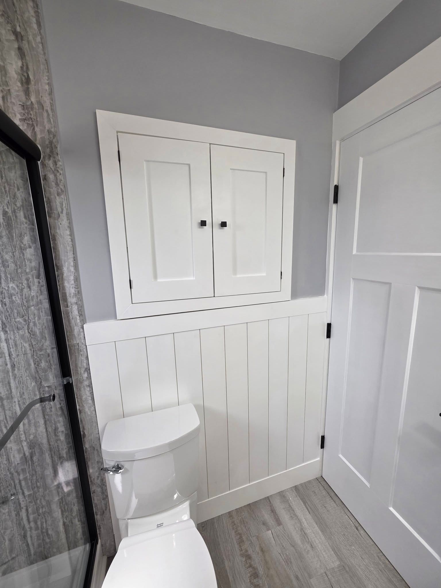 Bathroom with white wainscoting, toilet, storage cabinet, and gray walls; door on the right.