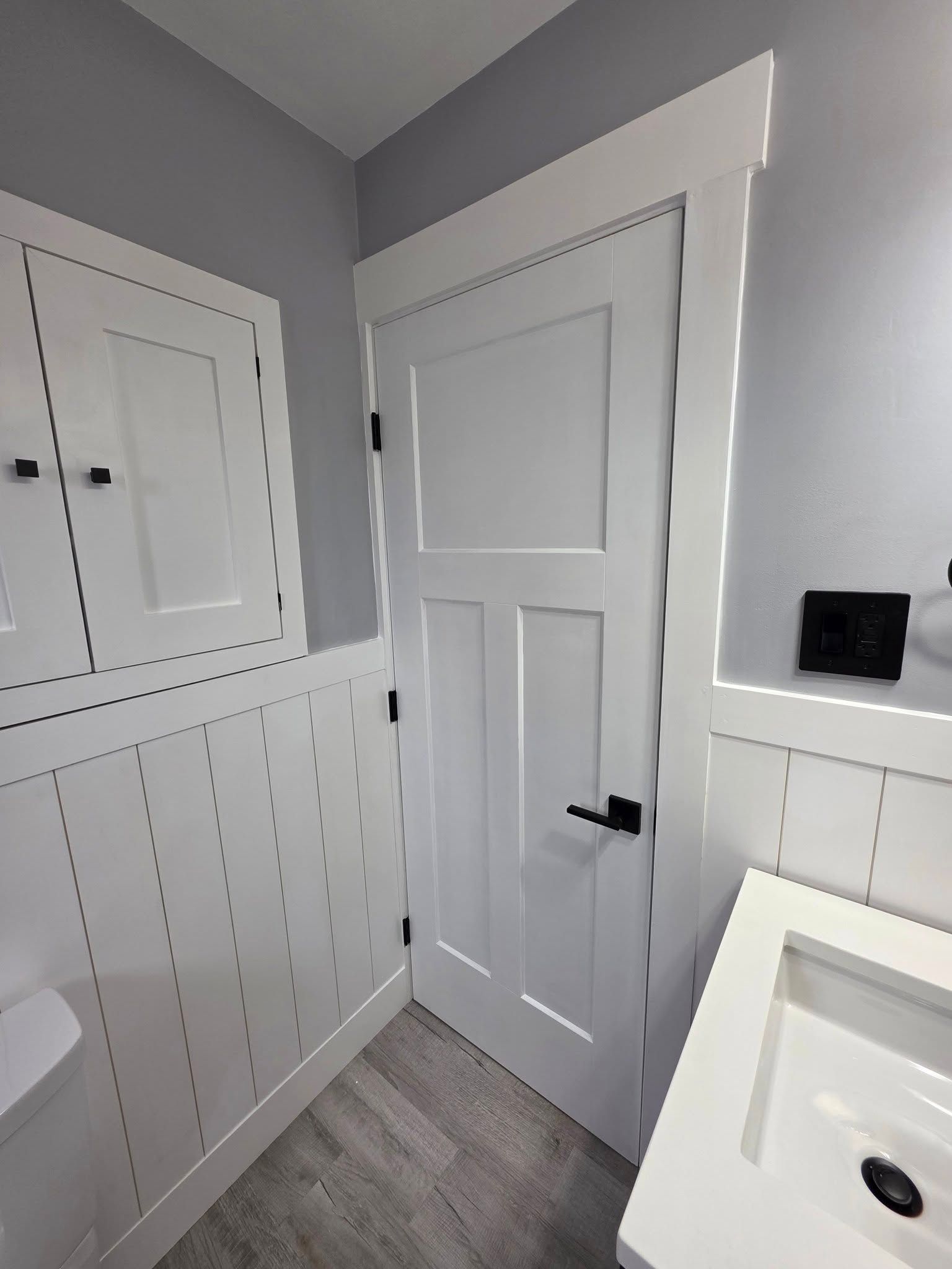 White bathroom with paneling, door, cabinet, and sink. Gray walls and floor.