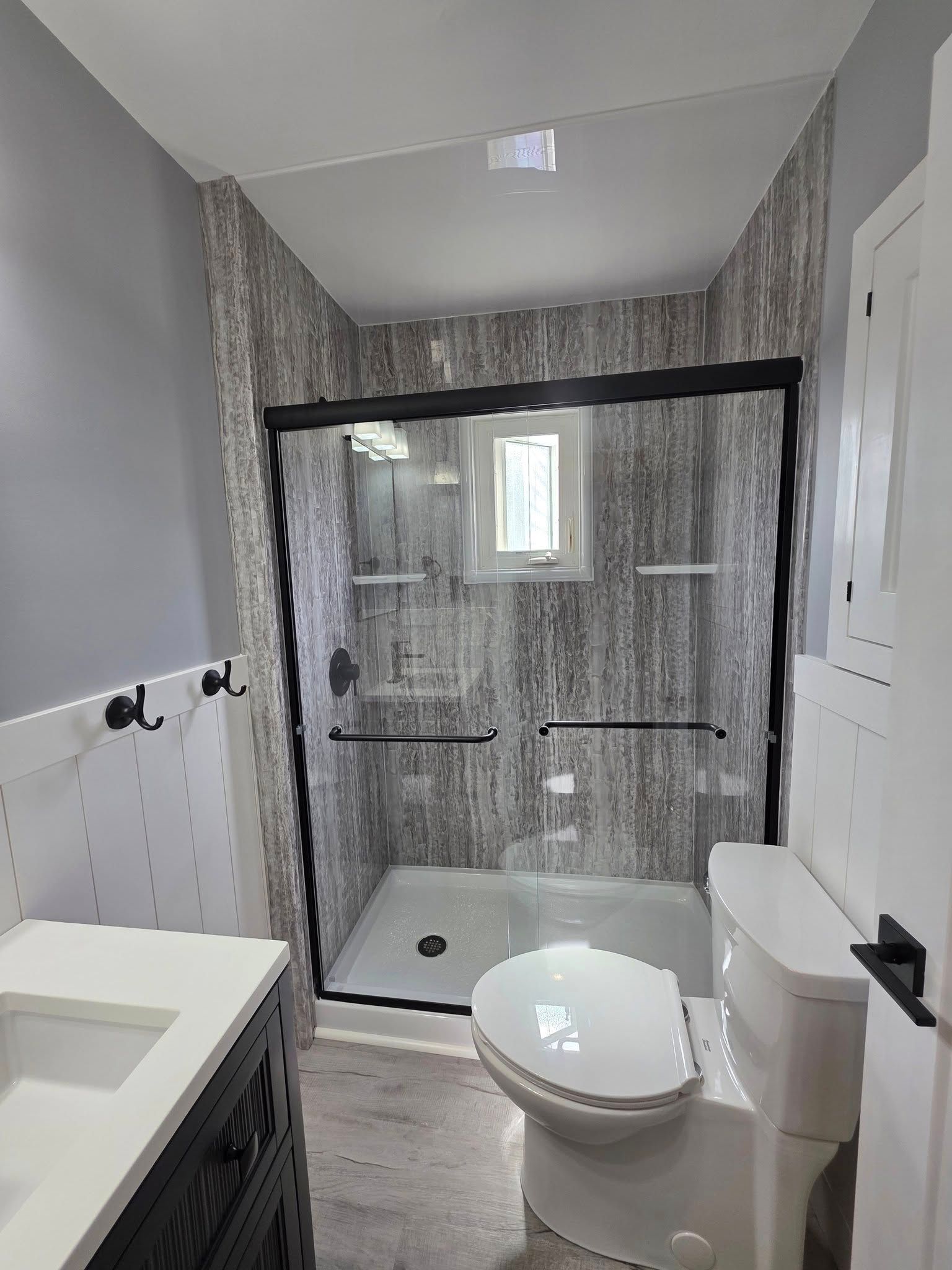 Bathroom with black-framed glass shower, patterned gray walls, white toilet, vanity, and cabinet.