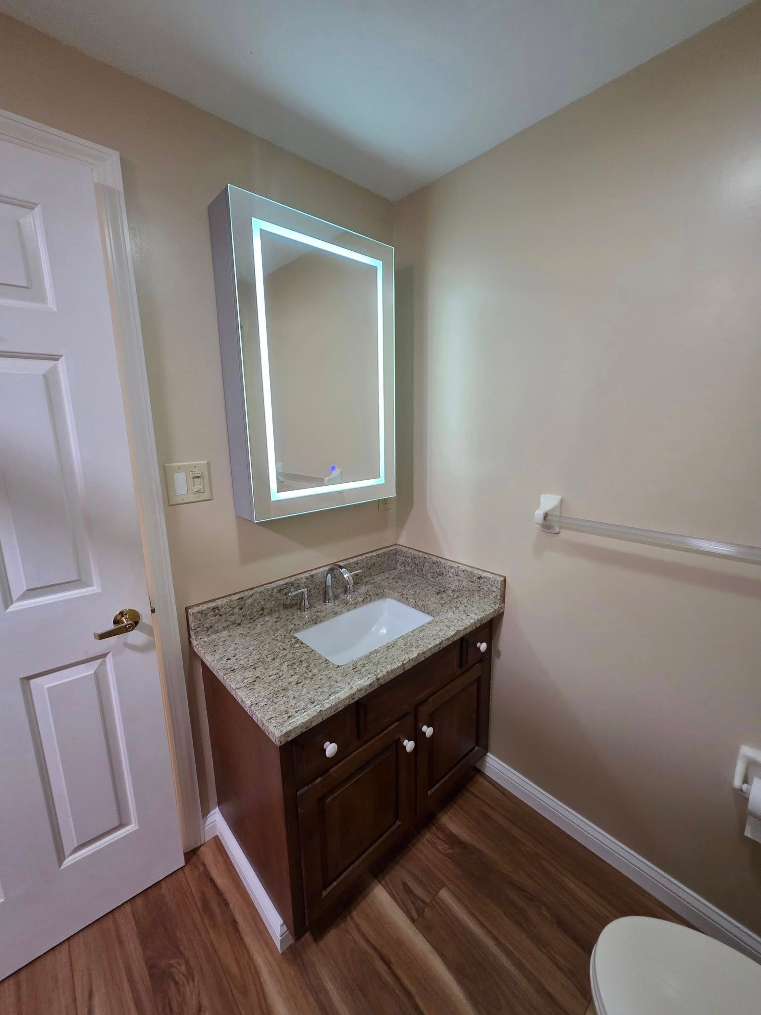 Bathroom with brown vanity, granite countertop, lit mirror, and wood-look floor. 