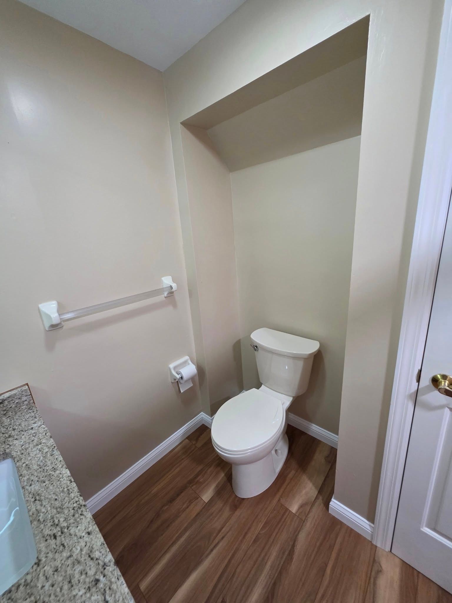 A small beige-walled bathroom with a toilet, towel rack, and wooden floor.