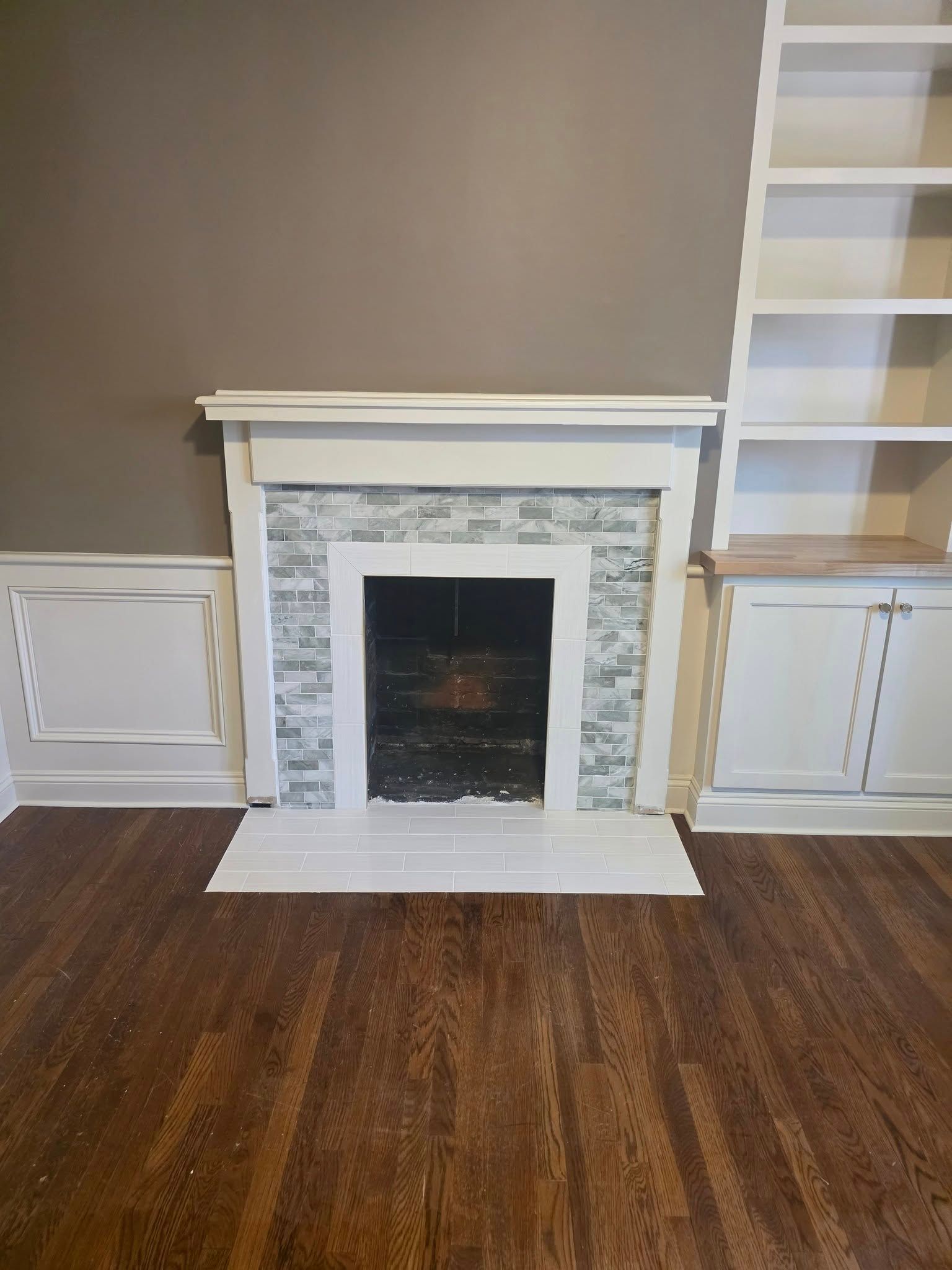 Fireplace with gray and white mosaic tile, white trim, and built-in bookshelves. Brown wooden floor and gray wall.