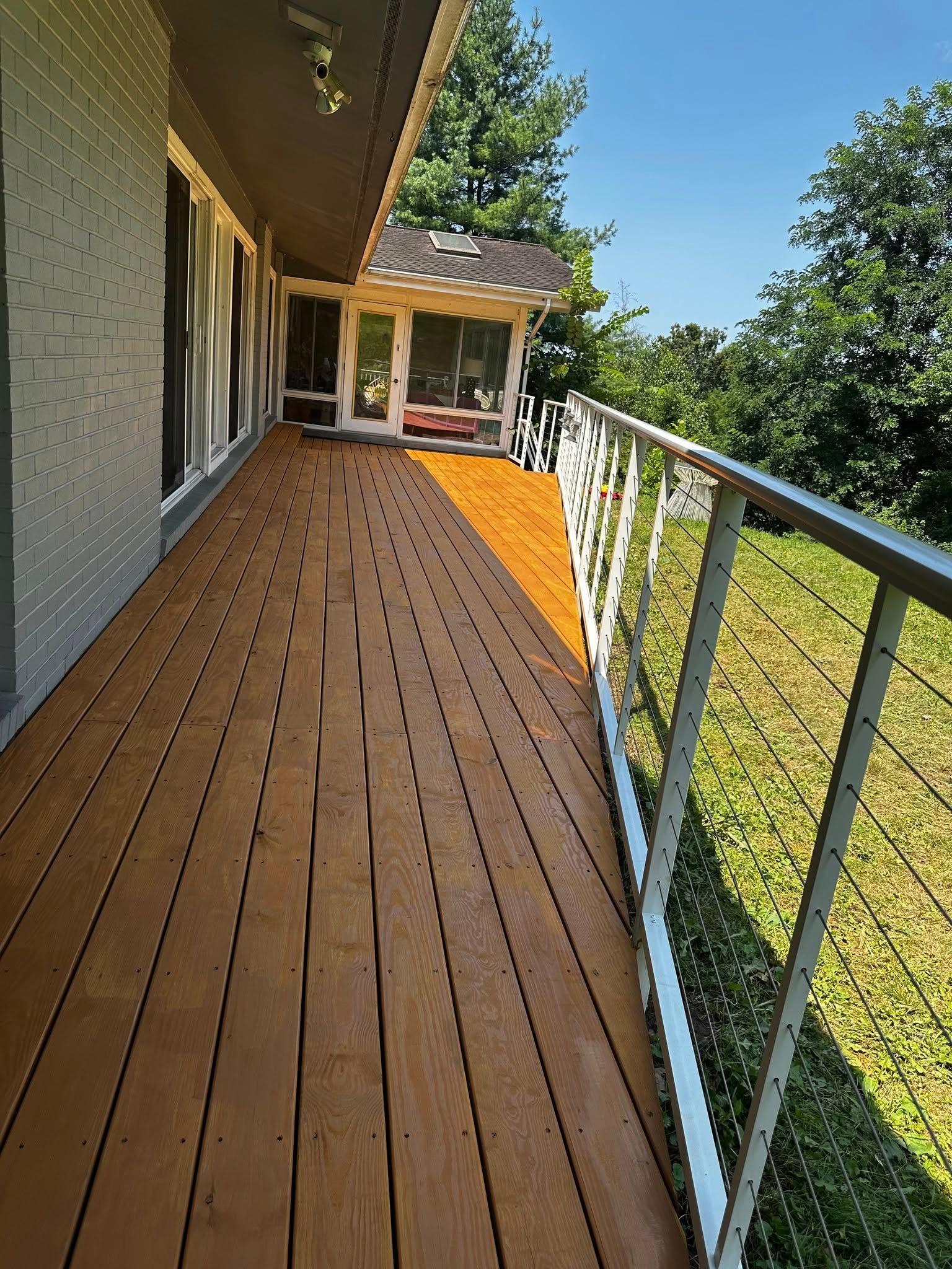 Wooden deck with metal railing, leading to a screened-in porch, set against a backdrop of greenery.