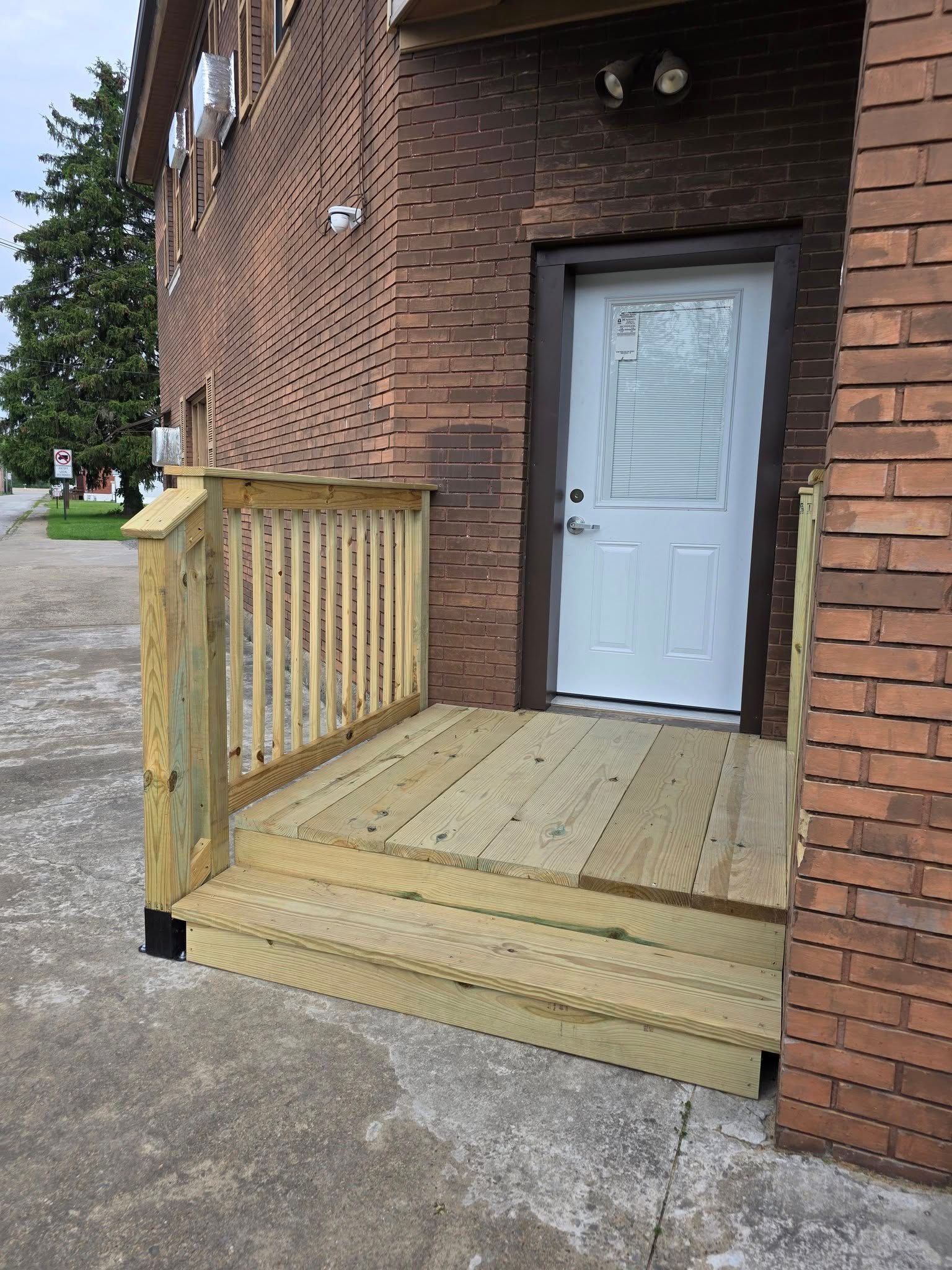 Wooden porch with handrails and steps leading to a white door on a brick building.