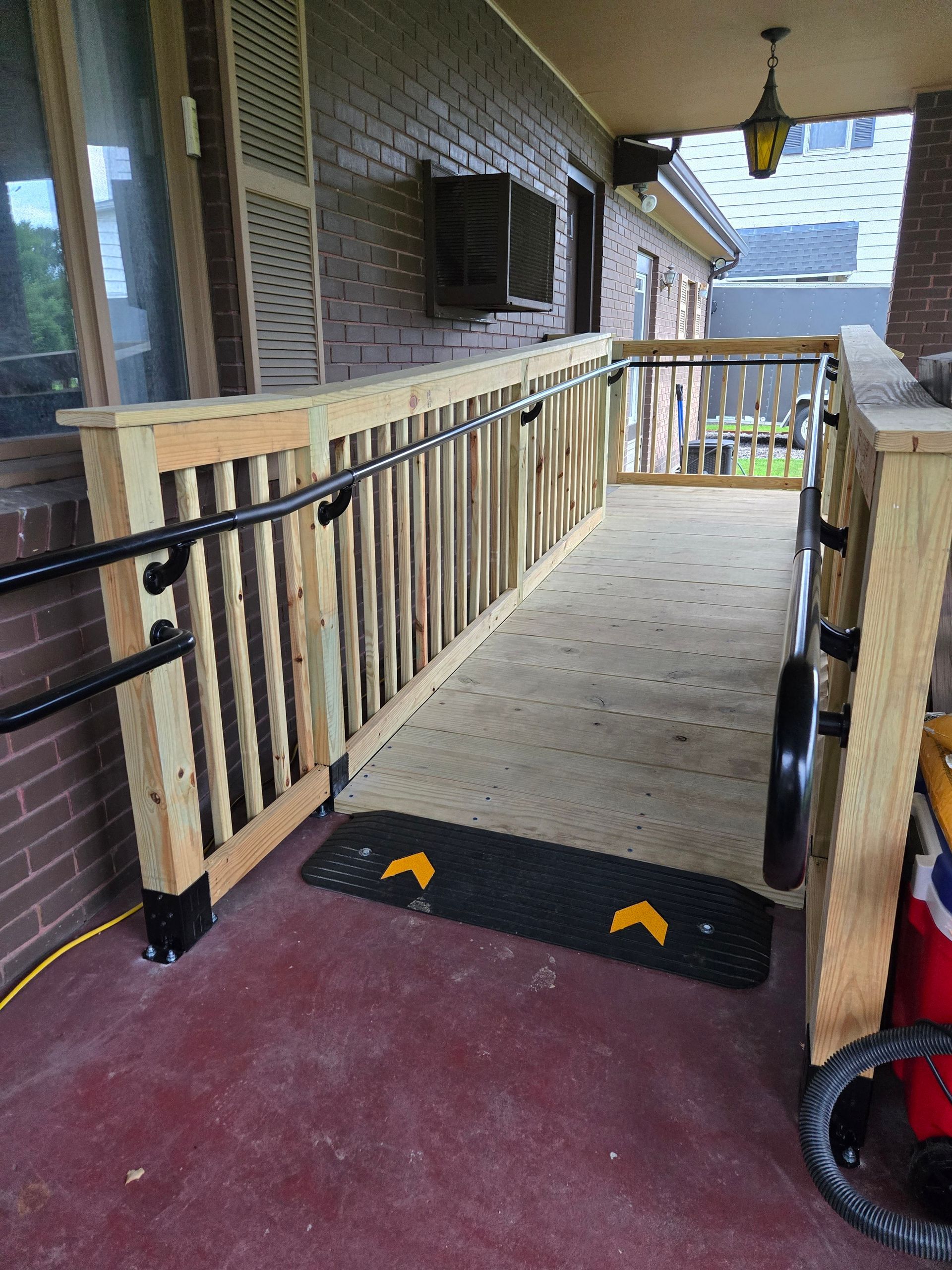 Ramp with wooden railing and black handrails on a brick porch, leading up to a house.