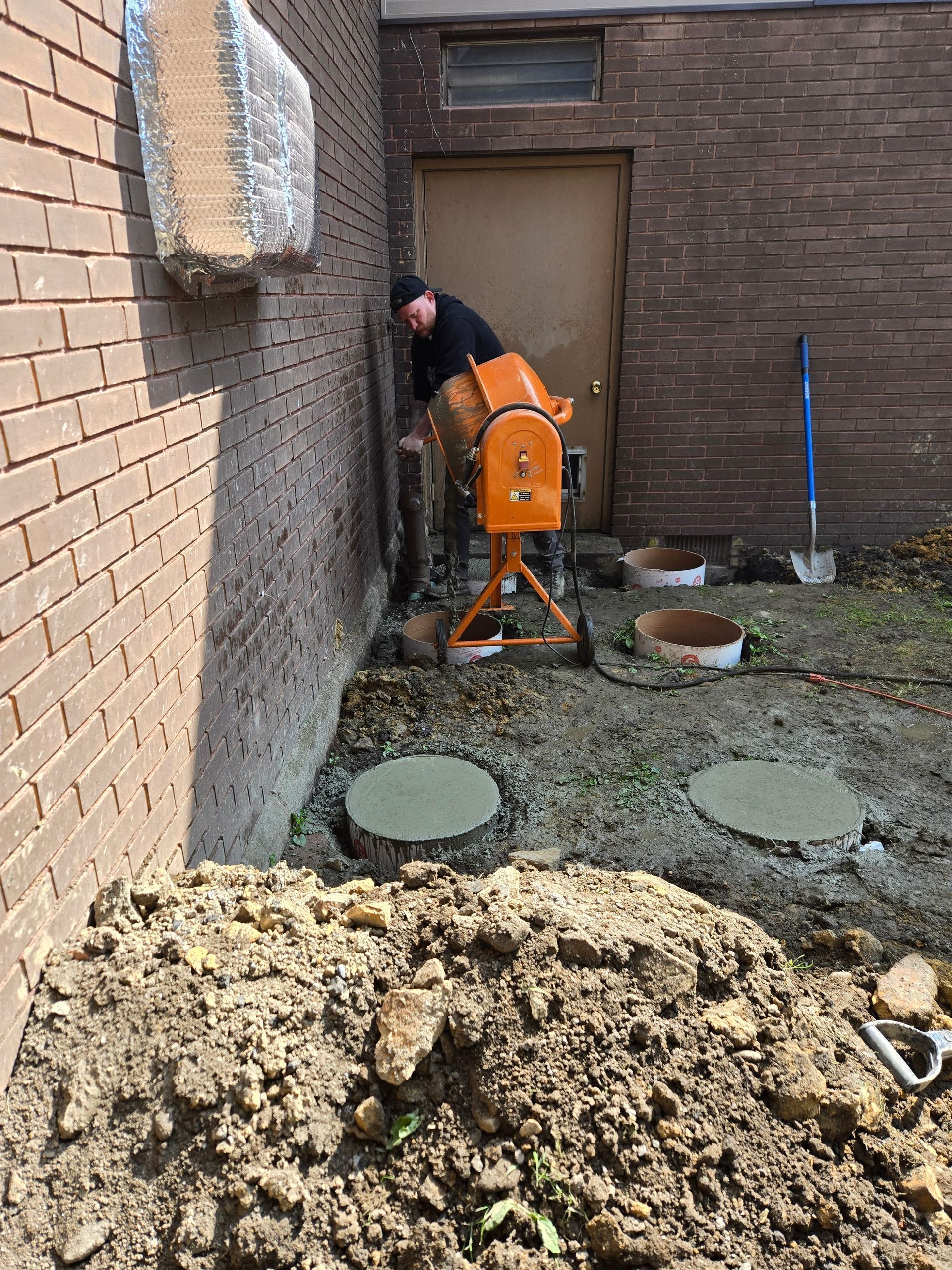 Person mixing concrete near a brick building with orange mixer. Soil and concrete covers visible.