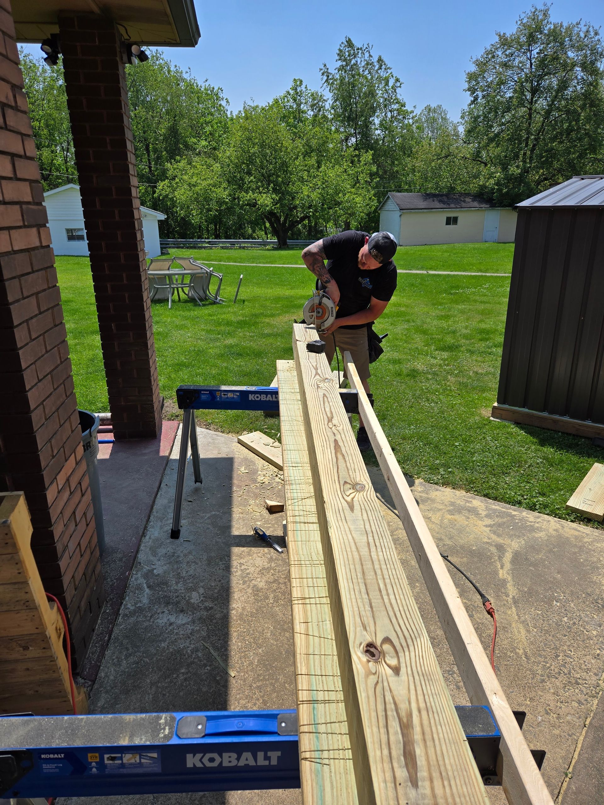 Man using a saw to cut lumber on a sawhorse in an outdoor setting. Sunny day.