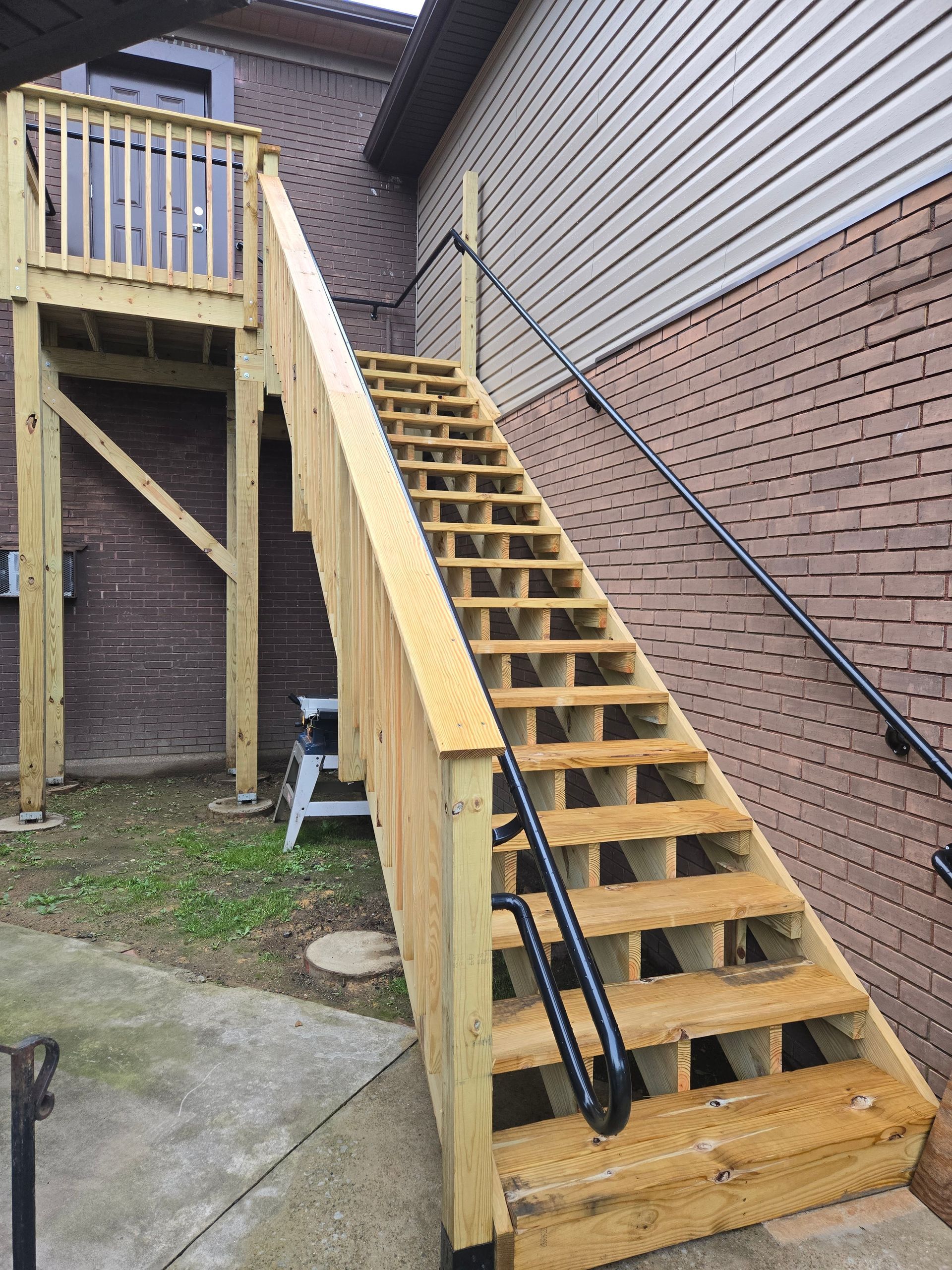 Wooden outdoor staircase leading to a raised deck, with black handrails on a brick and siding building.