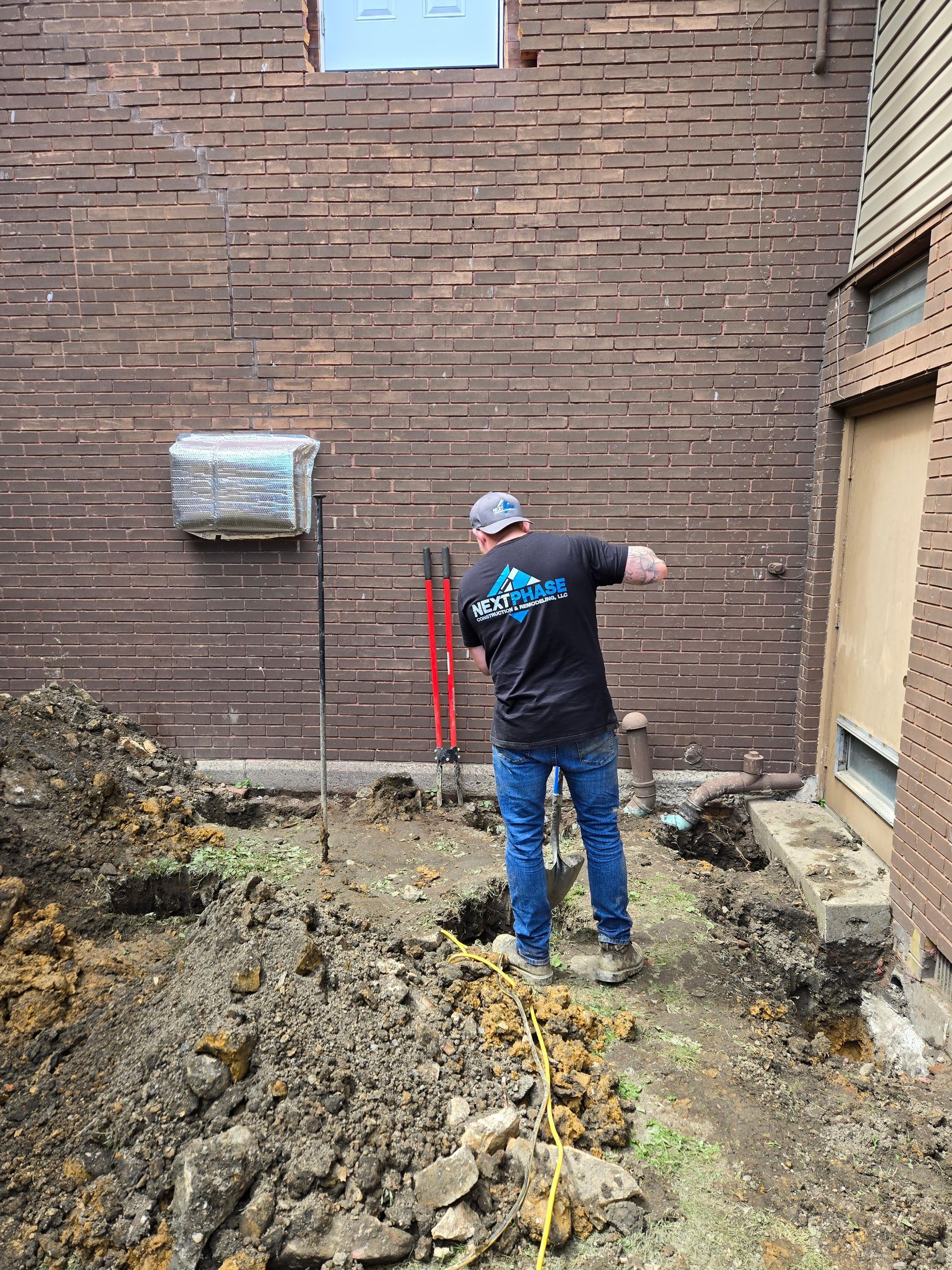 Man digging holes next to a brick building. Dirt and tools visible. Outdoors.