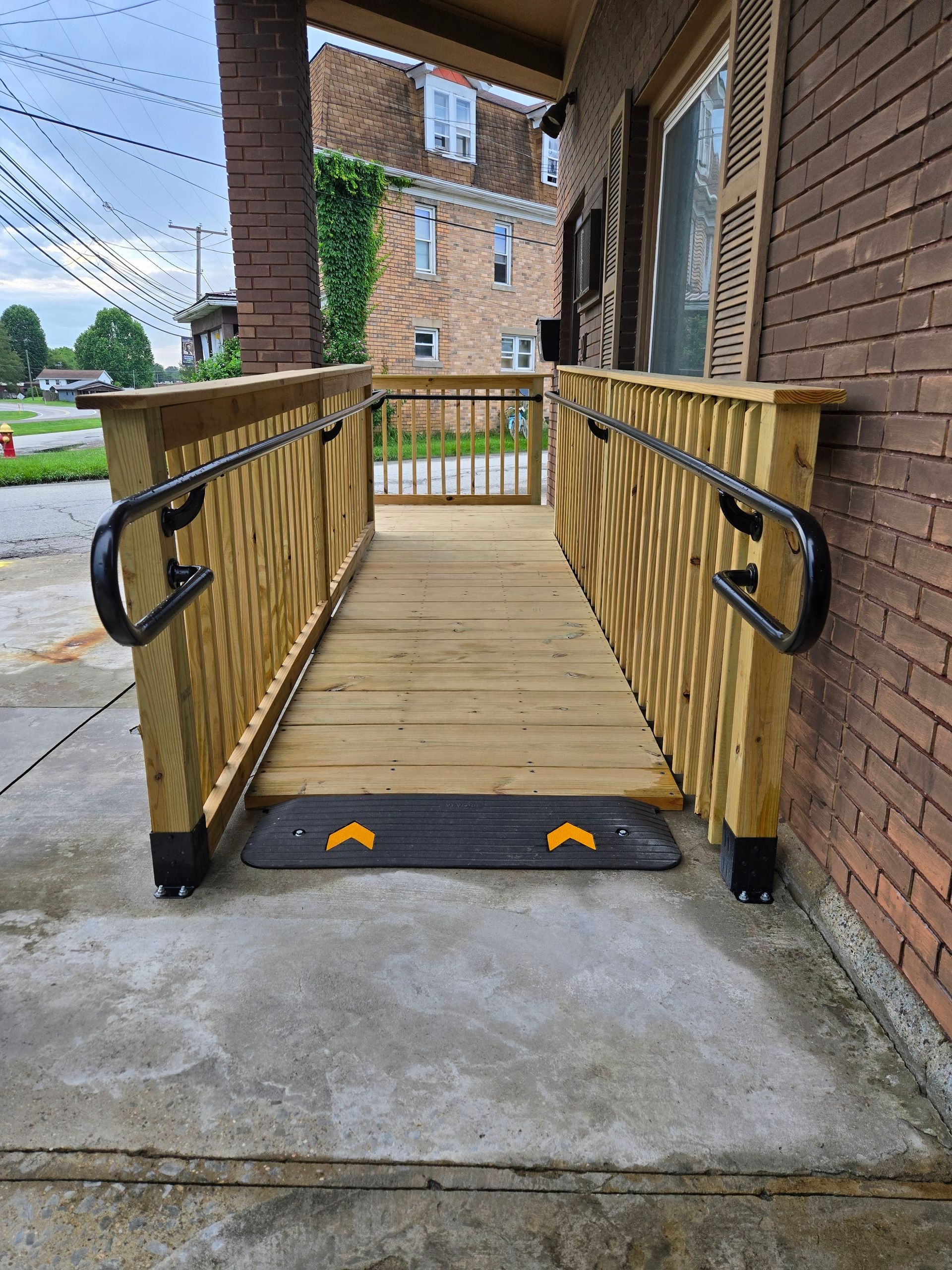 Wooden wheelchair ramp with black handrails on a brick building's porch.