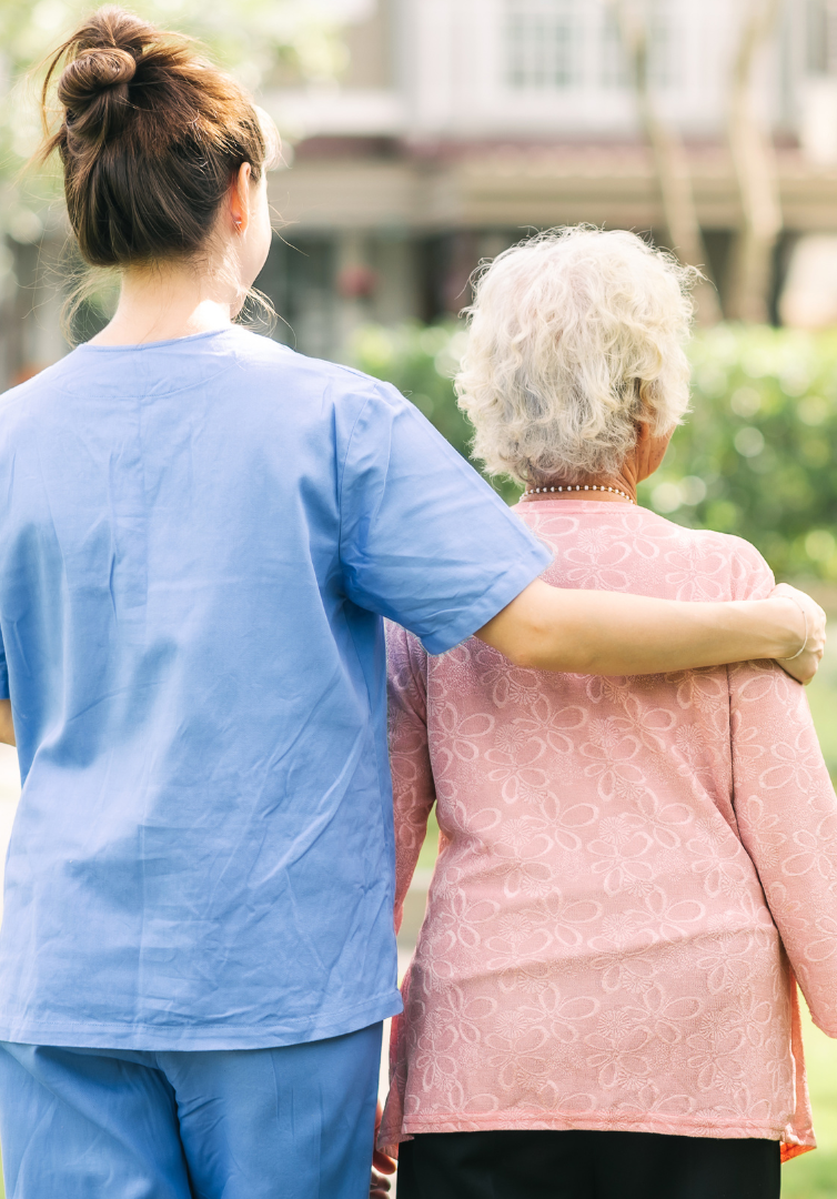 A caregiver in blue scrubs assists an elderly woman with white hair, arm around her shoulder, outdoors.