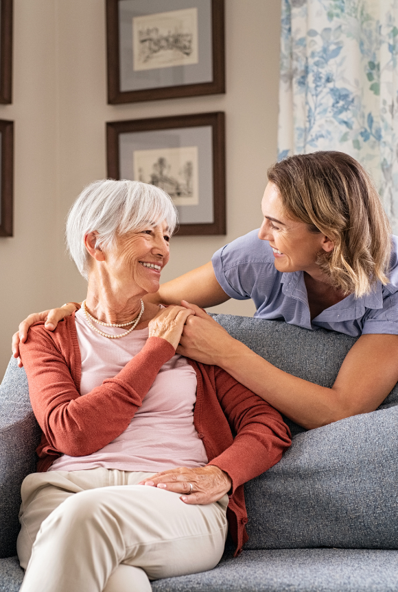 Woman with gray hair smiles as younger woman rests arm on her shoulder, both on couch.