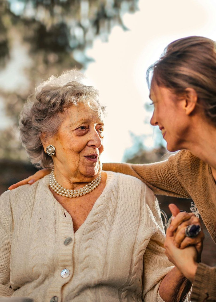 An elderly woman smiles, holding hands with a younger woman who has her arm around her.
