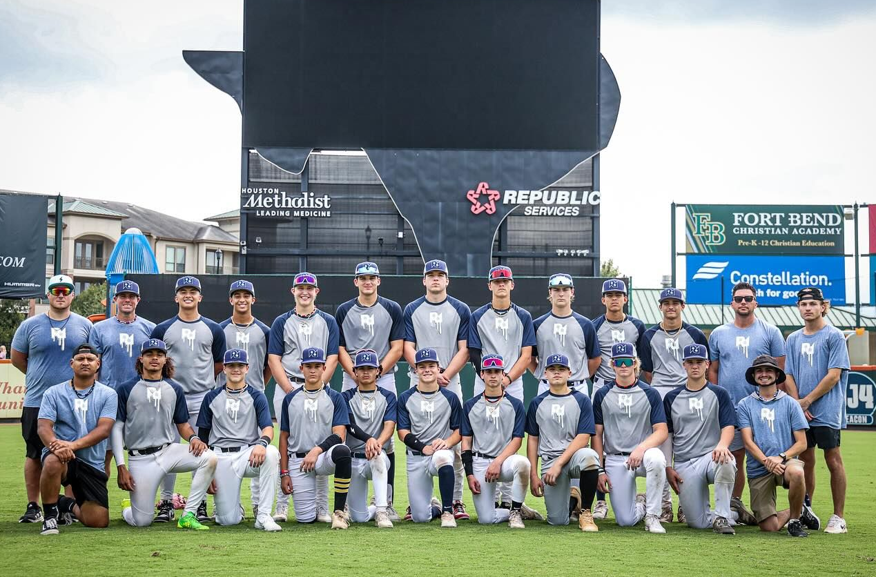 Baseball team in gray uniforms, posed on a green field in front of a scoreboard.