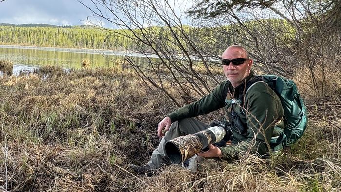 Man with camera sits in marshy grass by lake, wearing sunglasses, green outfit.