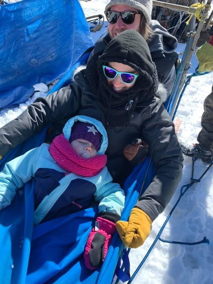 Two people in winter gear and sunglasses sit with a young child in a blue sled on a snowy surface.