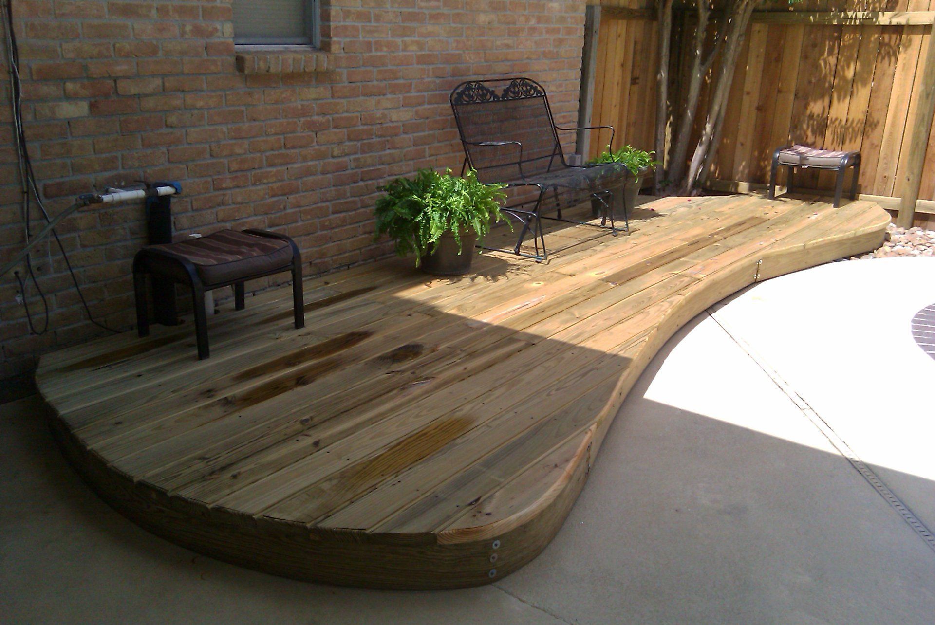 Wooden deck with curved edge, near brick wall and fence, holding furniture and plants.